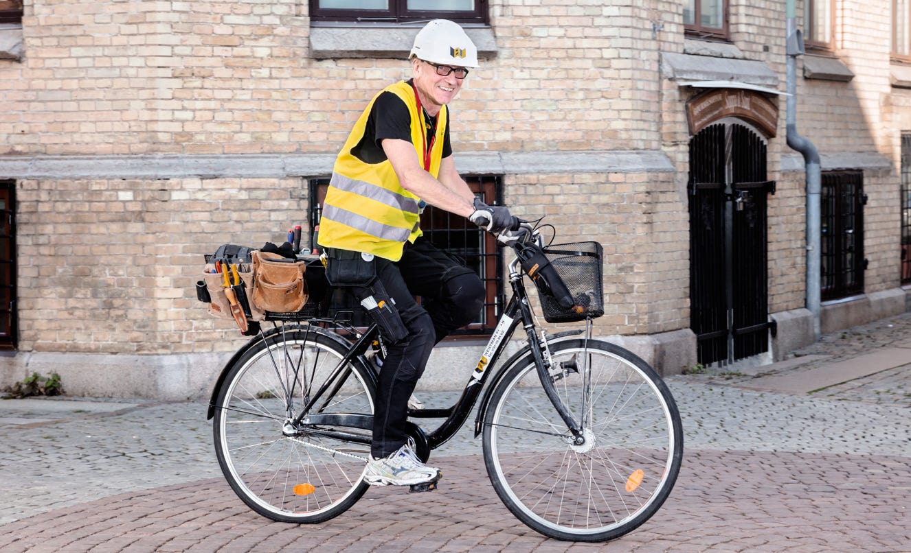 Person wearing a white helmet and yellow safety vest riding a bicycle on a cobblestone street with a brick building in the background.