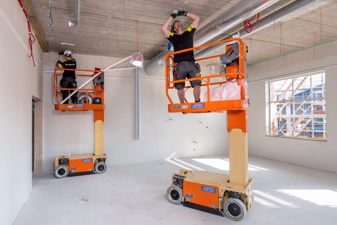 Two people on orange scissor lifts working on the ceiling of an unfinished room with exposed ductwork and large windows.