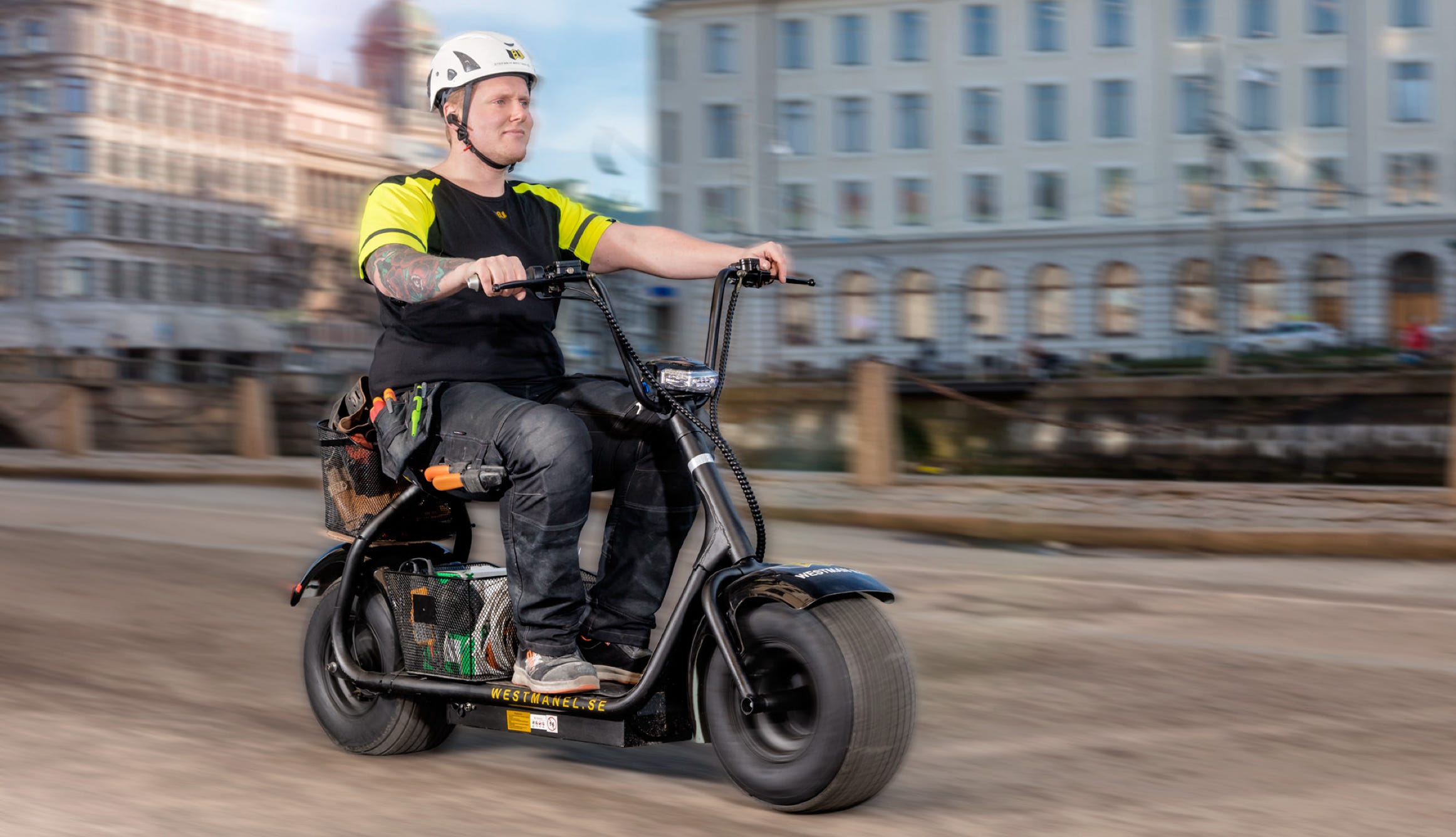 A person wearing a helmet and a black and yellow shirt rides a black electric scooter on a city street, with buildings in the background.