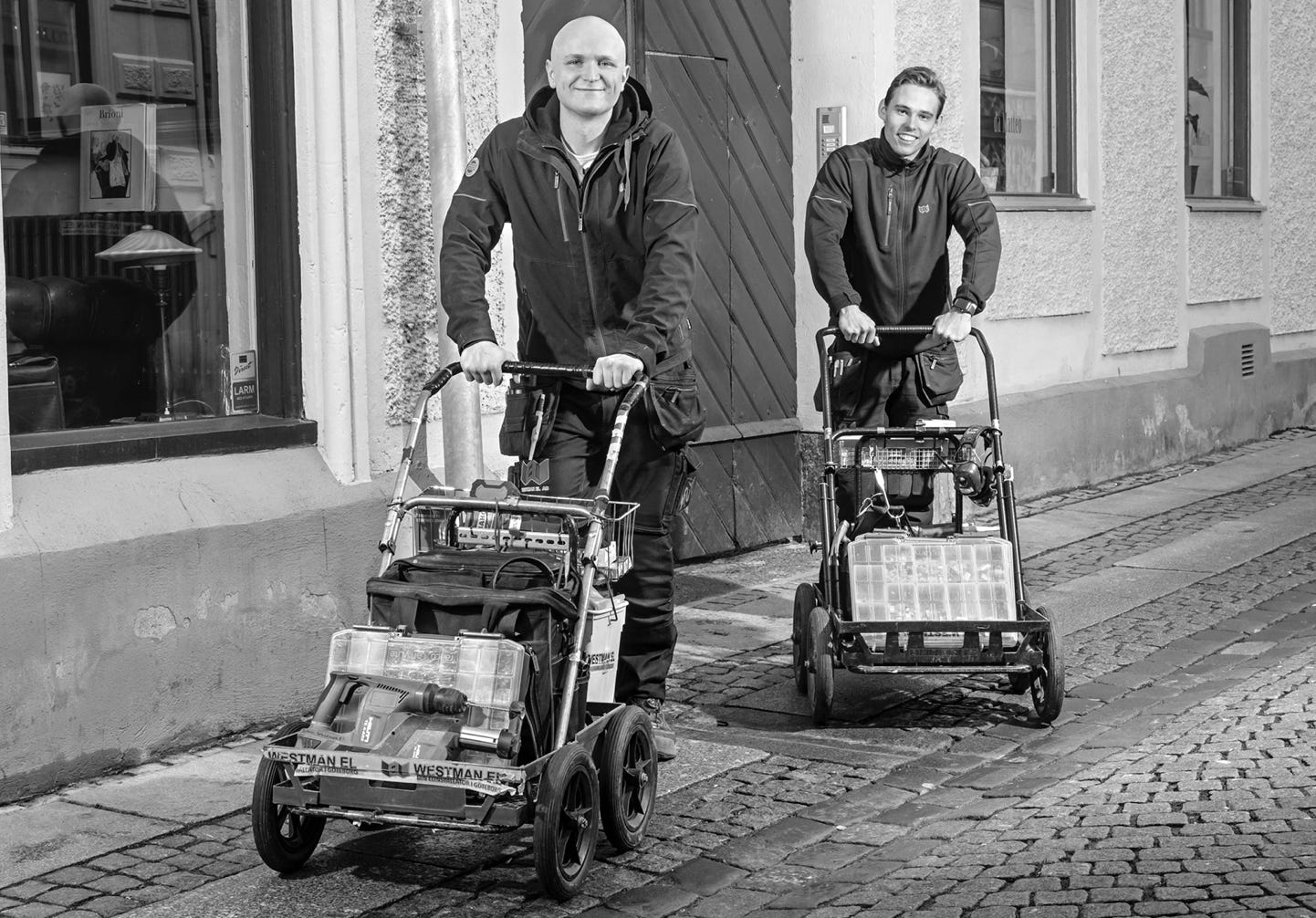Two people pushing carts filled with boxes on a cobblestone street, with buildings in the background. The image is in black and white.