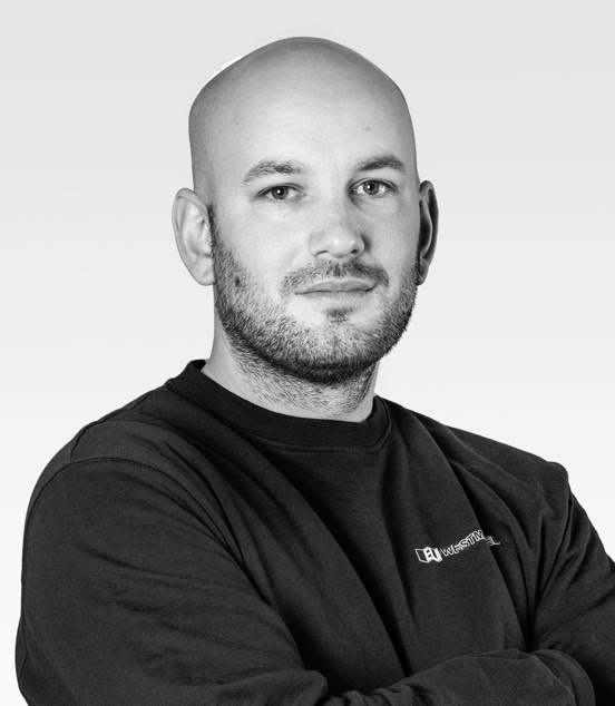 A person with a shaved head and a beard, wearing a dark long-sleeve shirt, standing with arms crossed against a light gray background.
