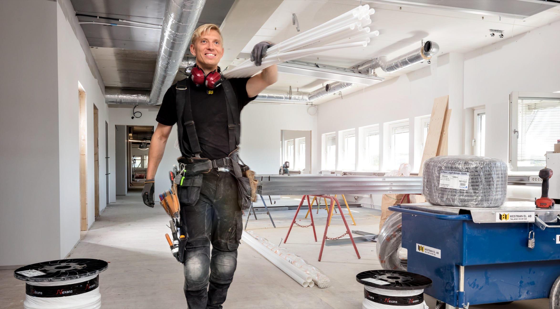 A person in work attire stands in a partially constructed room, holding a tool, with construction materials and equipment around.