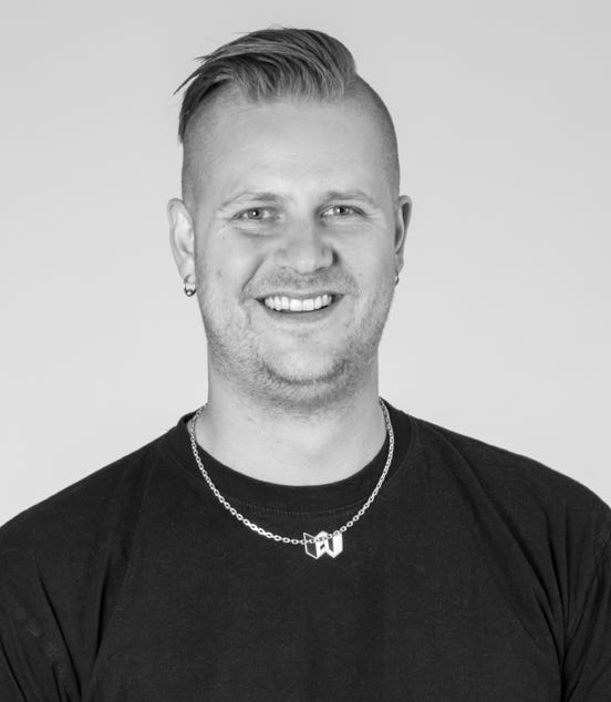 A person with short hair and a side shave, wearing a dark shirt and a necklace, smiling at the camera in a black and white photo.